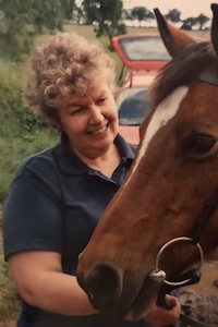 Maggie wearing a navy polo shirt whilst smiling and stroking a brown horse