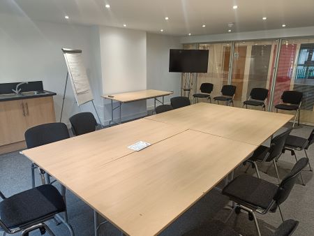 The meeting room is a medium-sized room with a large table in the centre surrounded by over ten chairs. There is a television or computer screen and sink. One wall is made of windows.