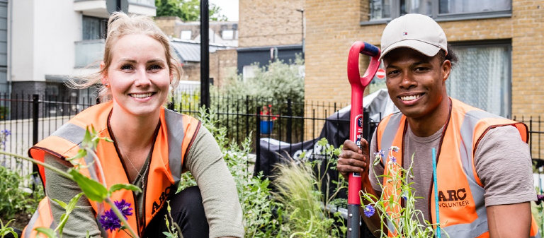 A man and woman in hi-visibility jackets and gardening tools in among some plants and smiling for the camera